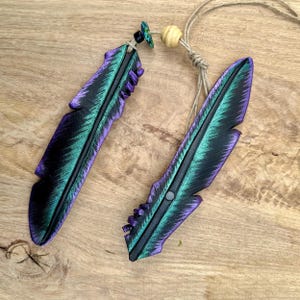 Peut inclure: Deux pendentifs en forme de plume présentant un dégradé de noir, de bleu sarcelle et de violet. Les pendentifs sont fixés à un cordon marron clair avec une petite perle en bois et une minuscule perle décorative bleue et verte.