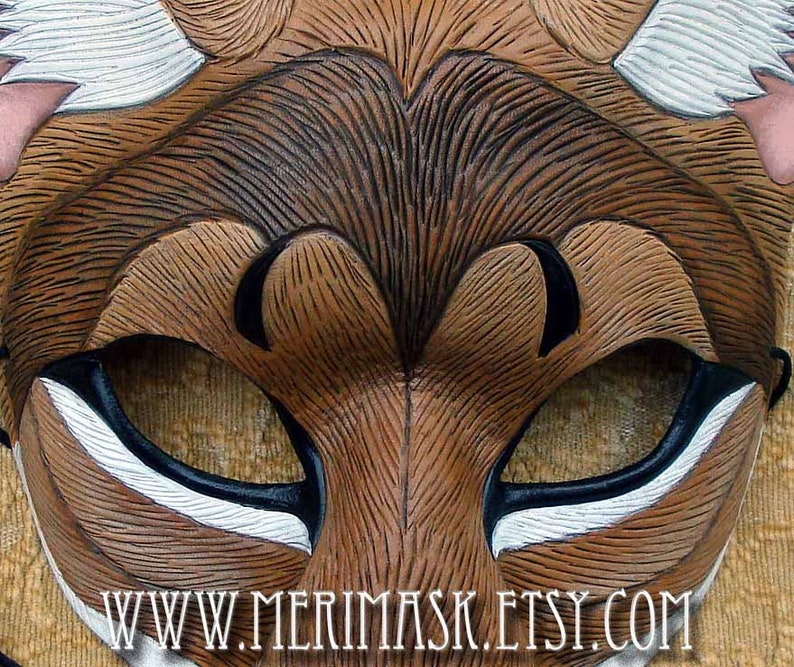 Puede incluir: Primer plano de una m&aacute;scara de cuero marr&oacute;n con detalles blancos y negros. La m&aacute;scara tiene un dise&ntilde;o de zorro con un patr&oacute;n de piel texturizado. La m&aacute;scara est&aacute; a la venta en el sitio web www.merimask.etsy.com.