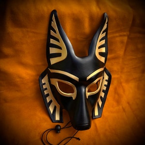 May include: A black leather mask with gold accents, shaped like an animal head with pointed ears. The mask has a detailed design with geometric patterns and a long snout.