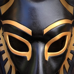 May include: A black leather mask with gold accents. The mask has two eye holes and a pointed nose.