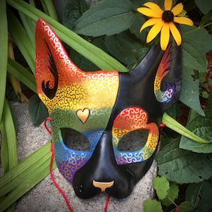 May include: A black cat mask with rainbow and gold swirls, a small heart, and black ears. The mask is on a green leafy background.