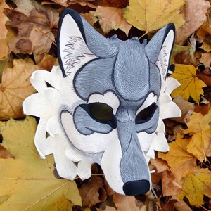 Op de afbeelding: Een handgemaakt wolfsmasker met een gedetailleerd ontwerp. Het masker heeft een grijs-wit kleurenschema met zwarte accenten rond de ogen en neus. Het masker ligt op een achtergrond van herfstbladeren.