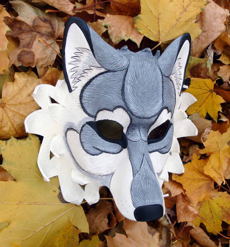 Op de afbeelding: Een handgemaakt wolfsmasker met een gedetailleerd ontwerp. Het masker heeft een grijs-wit kleurenschema met zwarte accenten rond de ogen en neus. Het masker ligt op een achtergrond van herfstbladeren.