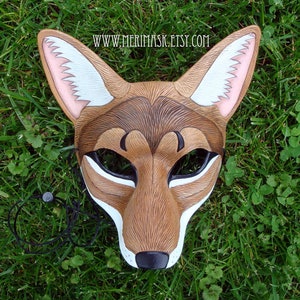 Puede incluir: Una m&aacute;scara de coyote de cuero marr&oacute;n con detalles de piel blanca y orejas rosas. La m&aacute;scara tiene ojos negros y una nariz negra. La m&aacute;scara est&aacute; sobre un fondo de hierba verde. www.merimask.etsy.com