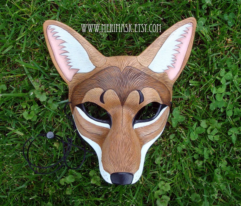 Puede incluir: Una m&aacute;scara de coyote de cuero marr&oacute;n con detalles de piel blanca y orejas rosas. La m&aacute;scara tiene ojos negros y una nariz negra. La m&aacute;scara est&aacute; sobre un fondo de hierba verde. www.merimask.etsy.com