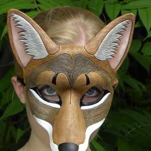 Puede incluir: Una m&aacute;scara de cuero marr&oacute;n y blanco con un dise&ntilde;o de lobo o coyote. La m&aacute;scara tiene un hocico largo, orejas puntiagudas y una nariz negra. La m&aacute;scara la lleva puesta una persona con el pelo rubio.