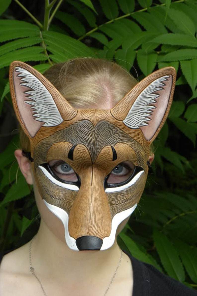 Puede incluir: Una m&aacute;scara de cuero marr&oacute;n y blanco con un dise&ntilde;o de lobo o coyote. La m&aacute;scara tiene un hocico largo, orejas puntiagudas y una nariz negra. La m&aacute;scara la lleva puesta una persona con el pelo rubio.