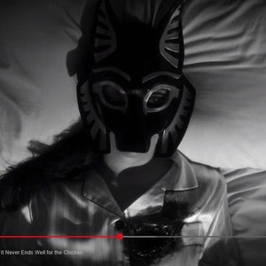 May include: A person wearing a black and silver animal mask lies in bed with white sheets. The mask has a pointed snout and large, round eyes. The person is wearing a white shirt with a black stain on the front. The text "Lucifer S5 E4 It Never Ends Well for the Chicken" is visible at the bottom of the image.