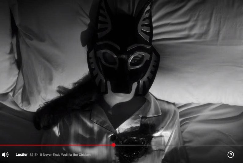 May include: A person wearing a black and silver animal mask lies in bed with white sheets. The mask has a pointed snout and large, round eyes. The person is wearing a white shirt with a black stain on the front. The text "Lucifer S5 E4 It Never Ends Well for the Chicken" is visible at the bottom of the image.