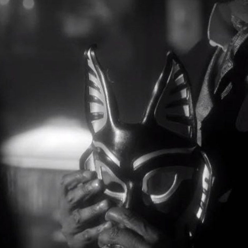 May include: A black and white mask with a cat-like design. The mask has large ears and a pointed nose. The eyes are outlined in white and the mask has a textured surface.