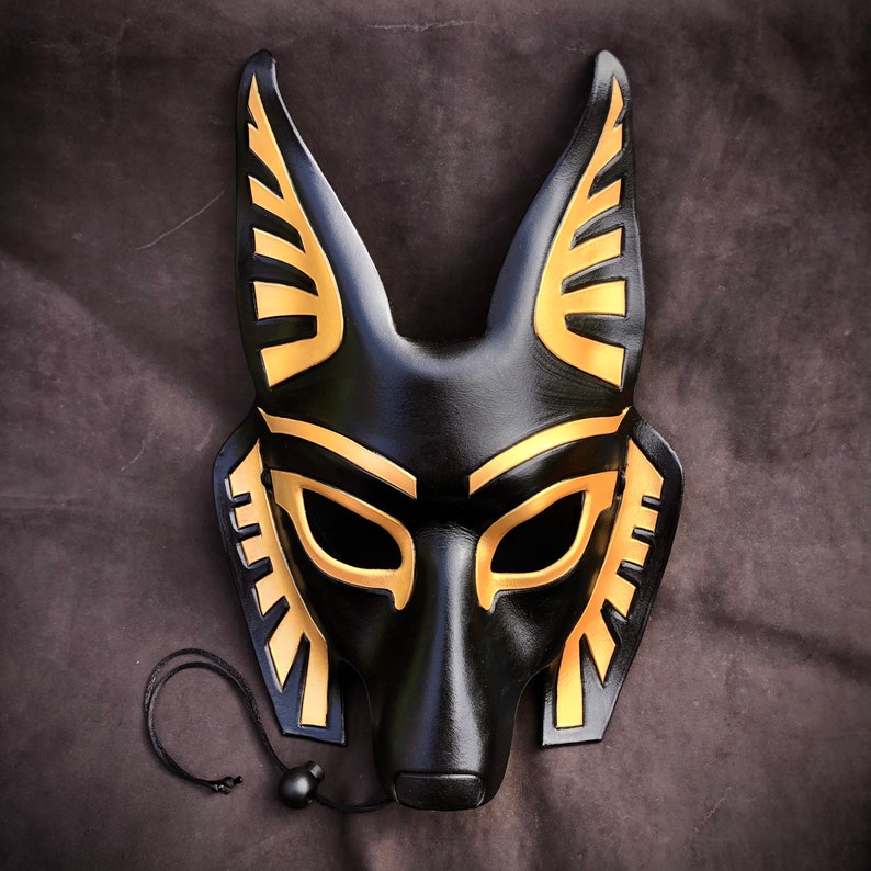 May include: A black leather mask with gold accents in the shape of an anubis jackal head. The mask has a long, pointed snout and large, almond-shaped eyes.
