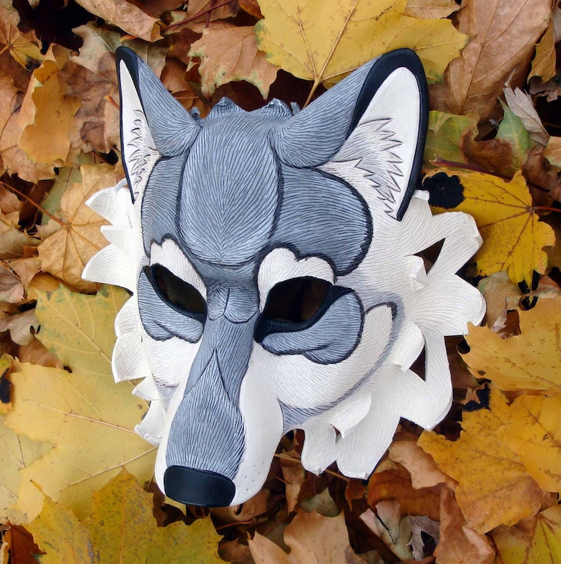 Op de afbeelding: Een handgemaakt wolfsmasker met een gedetailleerd ontwerp. Het masker heeft een grijs en wit kleurenschema met zwarte accenten rond de ogen en neus. De rand van het masker is voorzien van een witte, getextureerde vachtachtige rand. De achtergrond bestaat uit herfstbladeren.