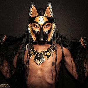 May include: A person wearing a black and gold mask with anubis-like features. The mask has a golden collar with an ankh symbol and a winged scarab beetle. The person has long black hair and is wearing black gloves.