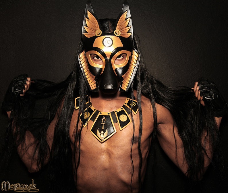 May include: A person wearing a black and gold mask with anubis-like features. The mask has a golden collar with an ankh symbol and a winged scarab beetle. The person has long black hair and is wearing black gloves.