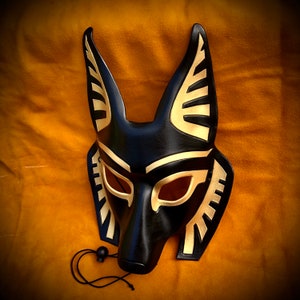 May include: A black leather mask with gold accents, resembling an animal face with pointed ears. The mask has a detailed design with geometric patterns and eye holes.