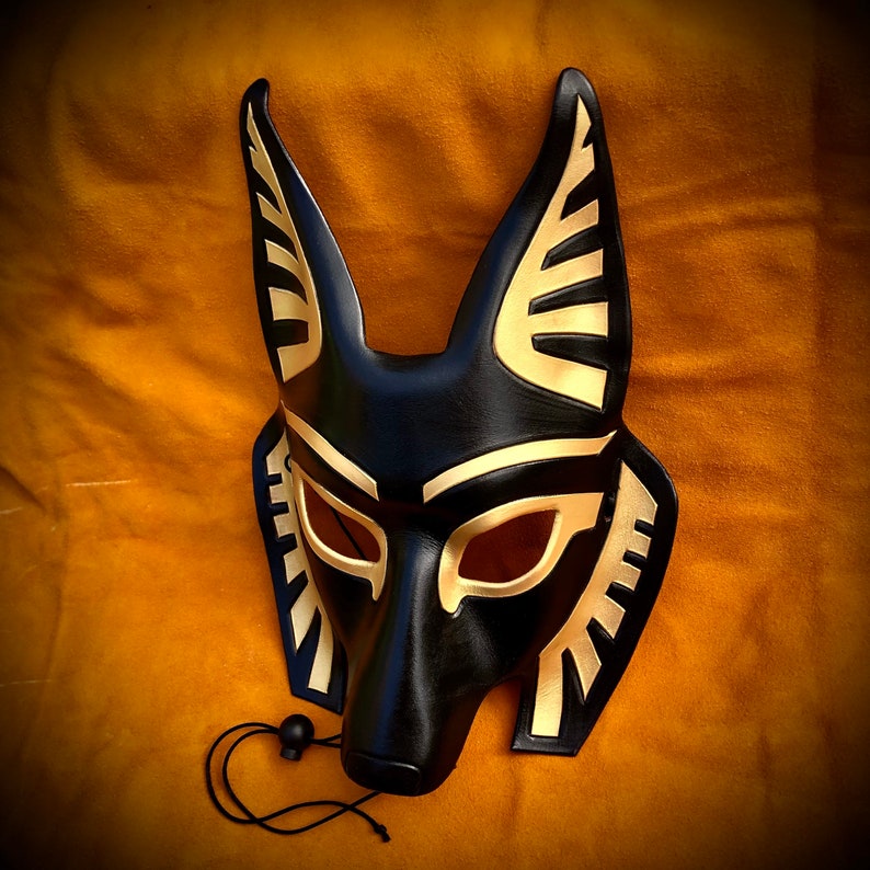 May include: A black leather mask with gold accents, resembling an animal face with pointed ears. The mask has a detailed design with geometric patterns and eye holes.