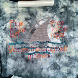 May include: A cross-stitch design on a gray and white tie-dye fabric. The design features a shark fin with the text "Fins to the left" and "Fins to the right" on either side of the fin. Below the fin is the text "You're the only bait in town".