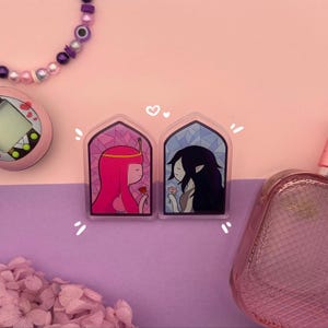May include: Two acrylic pins depicting cartoon characters in a stained-glass effect. One shows a pink-haired character with a rose, the other a black-haired character with flowers. Set against a pink and purple backdrop.