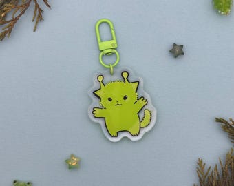 May include: A lime green alien-like character keychain with a matching green clasp. The cartoon alien has a smiling face and outstretched arms. The keychain is on a light blue background with decorative elements.