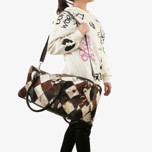 Puede incluir: Una bolsa de viaje de lona marrón y blanca con estampado de patchwork de cuero, con asas de cuero negro y una correa de hombro negra ajustable.