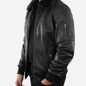 May include: A black leather bomber jacket with a brown fur collar. The jacket has a zipper closure, a small zippered pocket on the left sleeve, and ribbed cuffs and hem. The person is wearing black jeans.