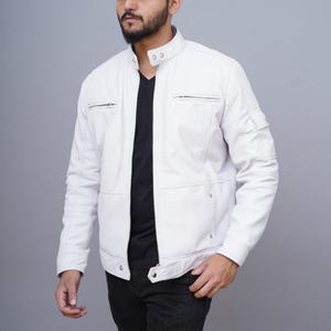 Könnte beinhalten: Eine weiße Lederjacke mit Stehkragen, Reißverschluss und mehreren Taschen. Die Jacke hat eine taillierte Passform und ein klassisches Design. Das Model trägt ein schwarzes Hemd und eine schwarze Hose.