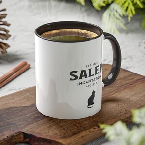 May include: White ceramic mug with a black handle and black text that reads "EST. 1626 SALEM INCANTATION SOCIETY" with a black silhouette of a cat. The mug is sitting on a wooden cutting board.