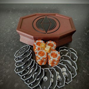 May include: A brown, geometric box with a black emblem, surrounded by stacks of orange and white game chips, black tokens, and dice. The game pieces are arranged on a dark surface, suggesting a board game.