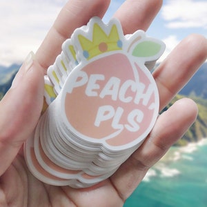 May include: A stack of peach-colored stickers with a crown and the text "PEACH PLS" in white. The stickers are in a hand against a blurred background of mountains and ocean.