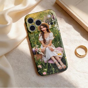 May include: Black phone case featuring a photograph of a person in a white dress and straw hat, sitting in a field of flowers. The case has a black border and cutouts for the camera lenses.