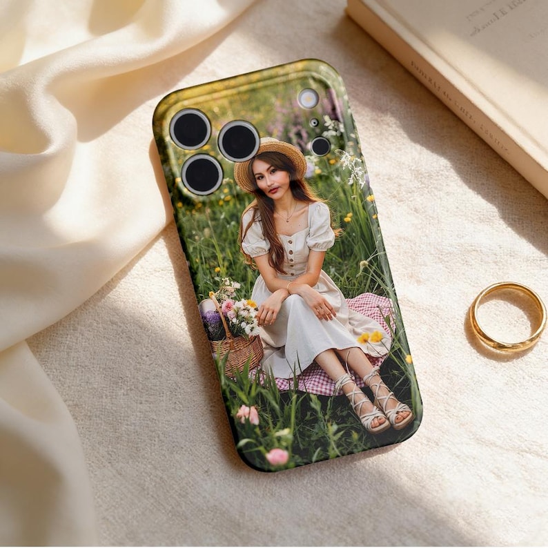 May include: Black phone case featuring a photograph of a person in a white dress and straw hat, sitting in a field of flowers. The case has a black border and cutouts for the camera lenses.