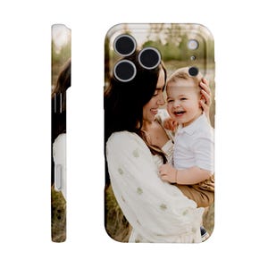 May include: A phone case featuring a photograph of a woman holding a smiling child. The case has a glossy finish and a protective design. The image on the case shows the woman in a white dress and the child in a white shirt and brown pants.