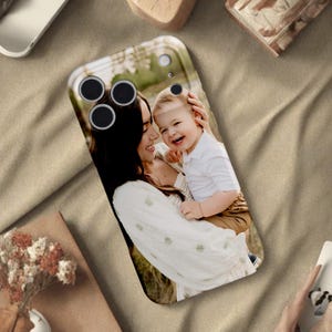 May include: A smartphone case featuring a photograph of a woman and a child. The case has a clear, glossy finish and a rectangular shape with rounded corners. The image shows a woman in a white dress holding a smiling child.