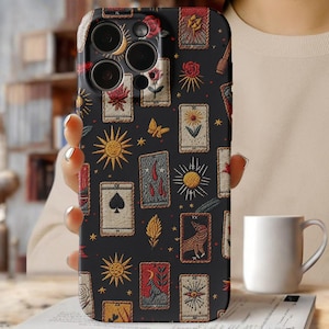 May include: Black phone case featuring a tarot card design with suns, moons, roses, and other symbols. The case has a dark background and a detailed, embroidered appearance. The case is held in a person's hand.