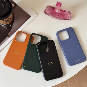 May include: Four phone cases in orange, dark green, black, and blue. Each case has a cutout for the camera and a gold monogram. A pink perfume bottle is in the background.