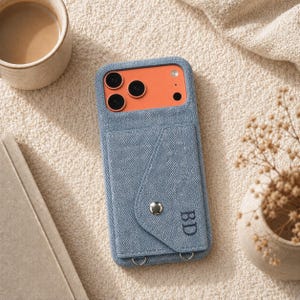 May include: A blue denim-textured phone case featuring an integrated card pocket with a silver snap button closure. The pocket is embroidered with the initials BD in dark thread. The case has a cutout revealing an orange phone back with three lenses.