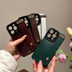 May include: A collection of phone cases in various colors, including burgundy, brown, white, and green. Each case features a textured, crocodile-like pattern, a black camera lens protector, and a wrist strap. The cases have gold-colored accents and a small, gold-colored button.