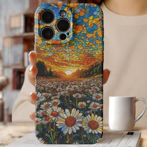May include: A phone case with a mosaic design depicting a sunset over a daisy field. The case shows a blue, yellow, and orange sky, and a field of white daisies with yellow centres. The case is held by a person.