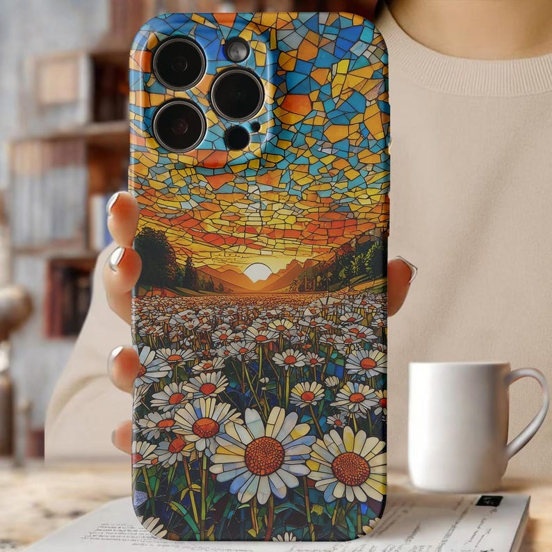 May include: A phone case with a mosaic design depicting a sunset over a daisy field. The case shows a blue, yellow, and orange sky, and a field of white daisies with yellow centres. The case is held by a person.