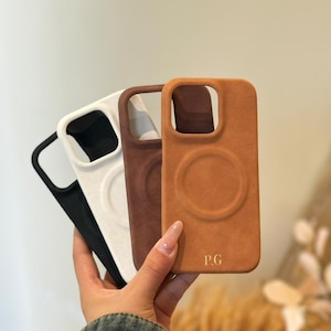 Pode incluir: Três capas para telemóvel em cores diferentes: preto, branco e castanho. A capa castanha tem um círculo elevado no centro e o texto "P.G." em letras douradas.