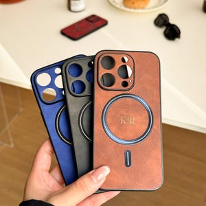 May include: Three phone cases with a built-in wireless charging pad. The cases are brown, black, and blue. The brown case has the initials "KR" on the charging pad.