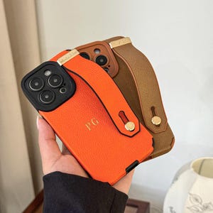 Peut inclure: Trois étuis de téléphone avec une texture en cuir et une sangle. Les étuis sont orange, marron et beige. L'étui orange porte les initiales "PG" en relief. Les étuis ont un accent en métal doré sur la sangle.