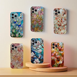 May include: A collection of six smartphone cases with stained glass floral designs. The cases feature various flower patterns in shades of blue, pink, orange, and yellow. The cases are displayed on a light brown surface.