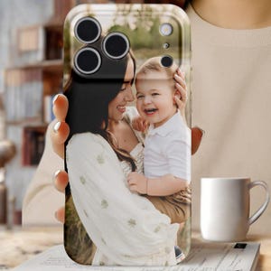 May include: A clear phone case featuring a photograph of a smiling woman and a child. The case has a black camera lens area and a glossy finish. The image on the case shows a woman in a white dress holding a laughing child.
