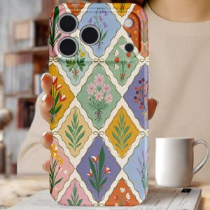 May include: A floral patterned phone case featuring a repeating diamond design with bouquets of flowers in various colors. The case has a colorful, vintage aesthetic, with a mix of pink, yellow, green, and purple hues. The phone case is held by a person.