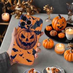 May include: Orange phone case featuring a jack-o'-lantern design with bats. The case is held in a hand, with other Halloween decorations like pumpkins and candles in the background.
