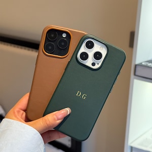 Può includere: Due custodie per telefono personalizzate. Una è color cuoio, l'altra verde scuro. La custodia verde ha le iniziali "D.G." in lettere dorate. Entrambe le custodie hanno un ritaglio per la fotocamera con più lenti.