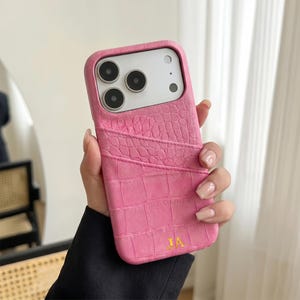 May include: A bright pink phone case with a textured crocodile skin pattern and a card slot pocket on the back. The case features the gold embossed initials JA at the bottom and has a cutout for a triple camera system on the upper left side.