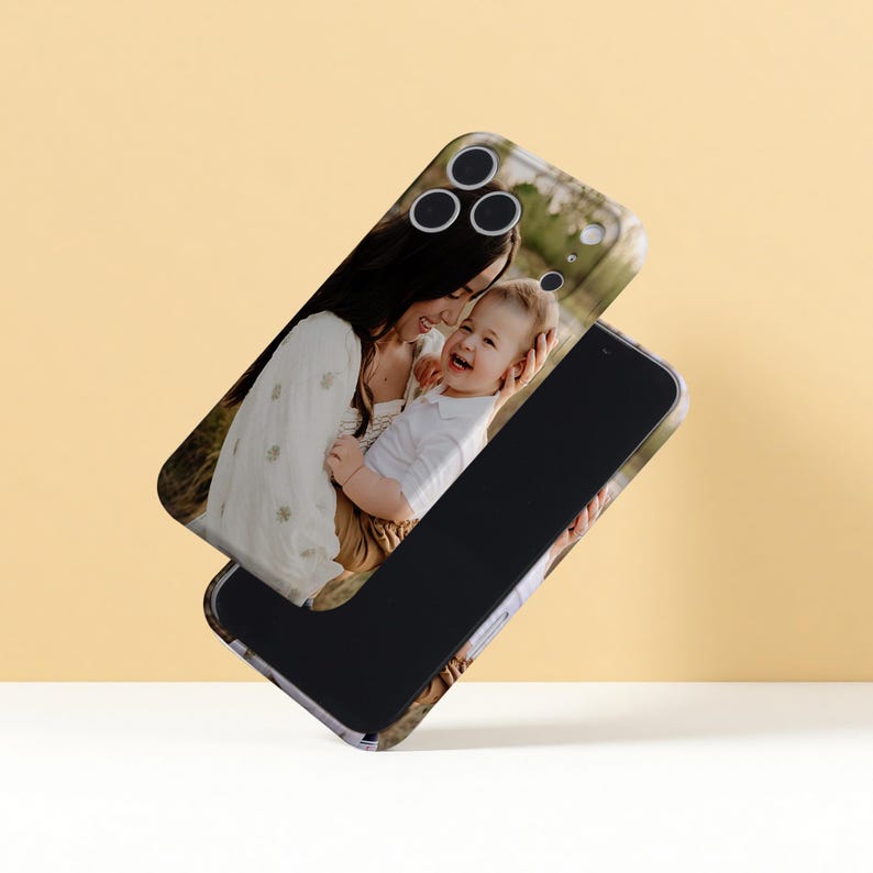 May include: Two phone cases, one with a photo of a smiling woman holding a baby, the other black. The top case shows the camera cutouts. The background is a soft yellow.