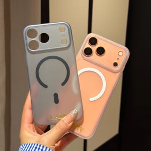May include: Two phone cases, one gray and one peach, are held in a person's hand. The gray case has a dark gray circular design and the word "LAM" in gold. The peach case has a white circular design.